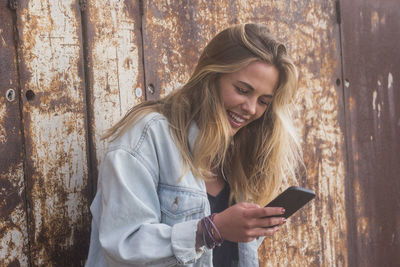 Smiling young woman using mobile phone against rusty metal