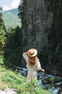 Young woman traveler with long blonde hair in straw hat looks at beautiful view of mountain river