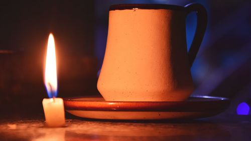 Close-up of illuminated candle by cup on table in darkroom