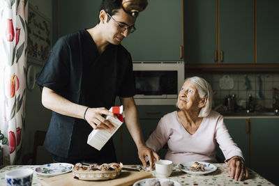 Male caregiver with juice box standing by senior woman in kitchen