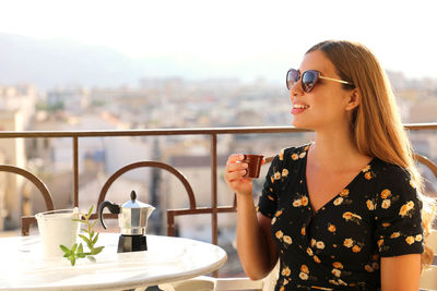 Young woman wearing sunglasses while standing by railing against cityscape