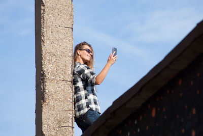 Low angle view of mid adult woman taking selfie with smart phone while standing against blue sky