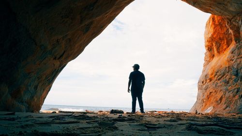 Rear view of man standing at beach against cloudy sky seen through cave