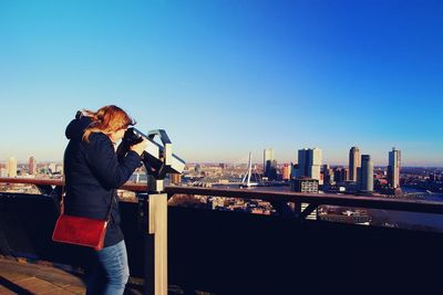 Woman looking at cityscape through coin-operated binoculars against blue sky
