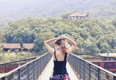 Woman tying hair while walking on footbridge against trees