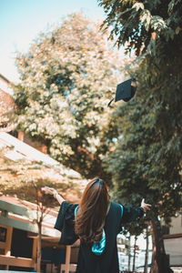 Rear view of woman catching mortarboard against trees
