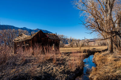 Scenic view of old wooden house along stream with bare winter trees against clear blue sky