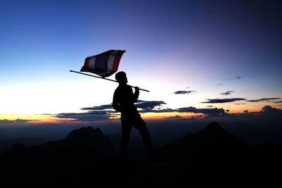 Silhouette person holding thai flag against sky during sunset