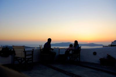 Silhouette people sitting by retaining wall against clear sky during sunset