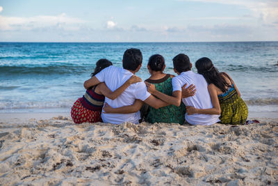 Rear view of friends sitting on beach against sky