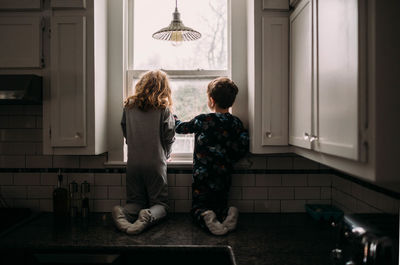 Brother and sister looking outside kitchen window on cloudy day