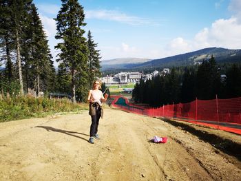 Full length of mature woman walking on dirt road against sky during sunny day