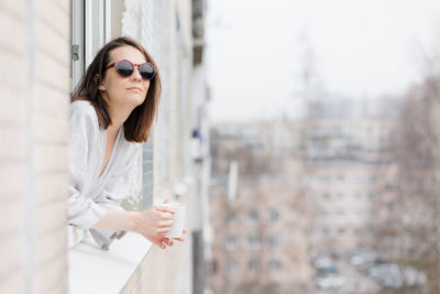 Portrait of woman wearing sunglasses while standing against window