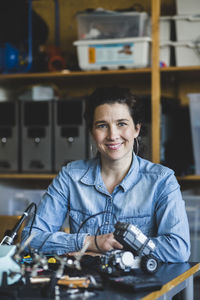 Portrait of confident mature female teacher sitting with science project at desk in classroom at high school