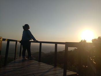 Rear view of man standing on railing against sky during sunset