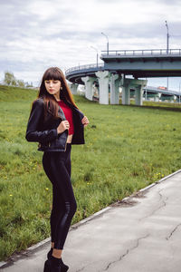 Young woman standing on footpath by field with bridge in background