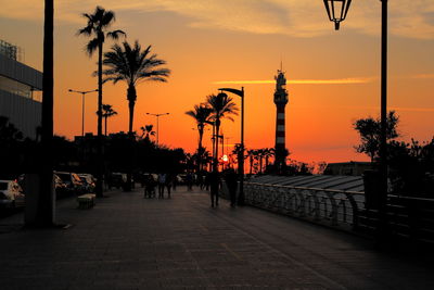 Silhouette people on street by palm trees against orange sky