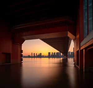 Bridge over river by buildings against sky during sunset