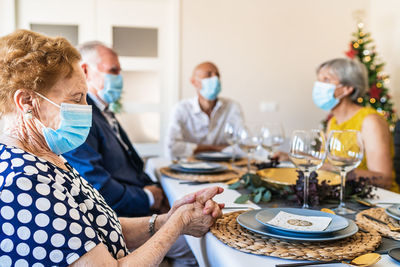 Positive senior friends in smart casual wear and face masks gathering together at dining table and having conversation at christmas eve