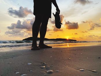 Low section of man standing at beach against sky during sunset