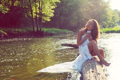 Young woman sitting by lake against trees