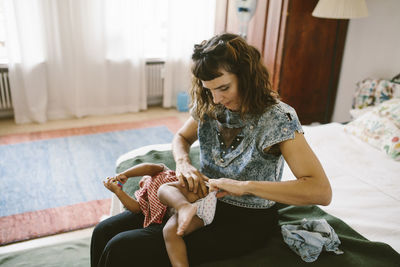 Mother adjusting diaper of daughter while sitting on bed at home