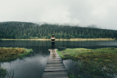 Rear view of woman standing on jetty in lake against sky during rainfall