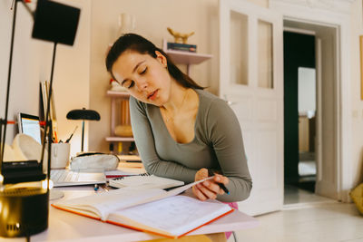 Young woman reading book while doing homework at table