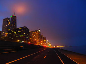 Illuminated road amidst buildings against sky at night