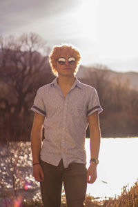 Portrait of young man in sunglasses standing at lakeshore against sky