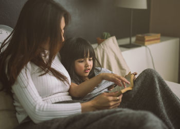 Mother reading book for daughter on bed at home