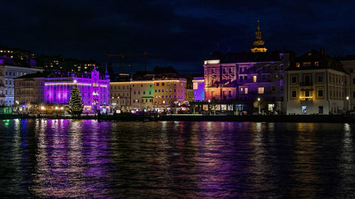 Illuminated buildings by river against sky at night