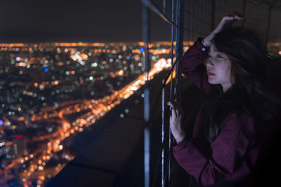 Woman looking illuminated cityscape through fence at night