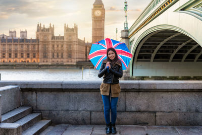 Portrait of young woman holding umbrella while standing against thames river in city