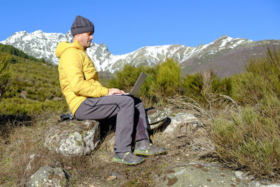 A man works with his computer in the wilderness near snow-capped mountains