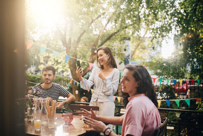 Smiling young woman video conferencing through smart phone while friends sitting in balcony during party