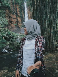 Woman holding hands of man standing against waterfall in forest