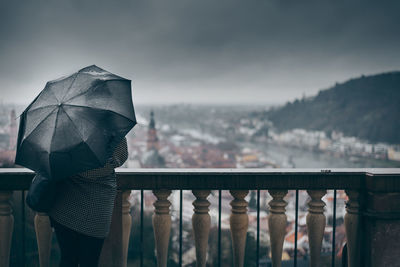 Rear view of woman with umbrella standing by railing against cloudy sky