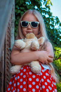 Beautiful happy girl in red polka dot dress soft toy on wooden balcony. cute joyful child long hair.