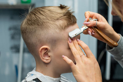 A boy is sitting in a barbershop, doing his hair with a sharp razor for a haircut