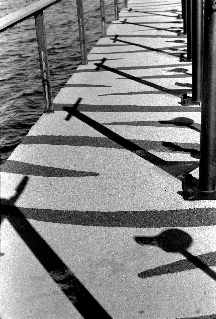 shadow, sunlight, black and white, line, day, black, nature, monochrome photography, monochrome, high angle view, footpath, the way forward, no people, white, railing, outdoors, iron, water, sunny, transportation, wood, walkway, architecture, focus on shadow, road, lane