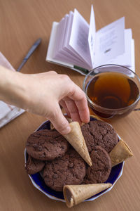 A woman's hand takes a waffle tube from a saucer with sweets, next to a cup of tea and an open book.