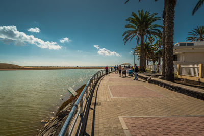 People walking footpath by sea against blue sky