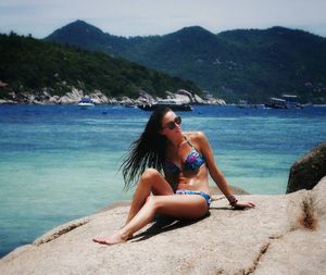 Woman wearing bikini sitting on rock against sea