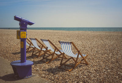 Telescope by deck chairs at beach against sky