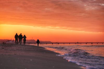 Silhouette people walking on shore at beach against orange sky