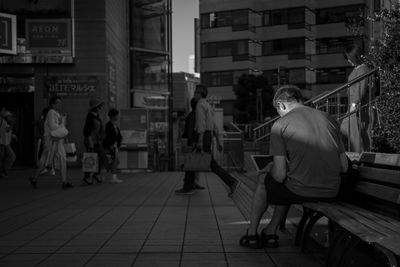 Rear view of man using laptop on bench near people walking on footpath