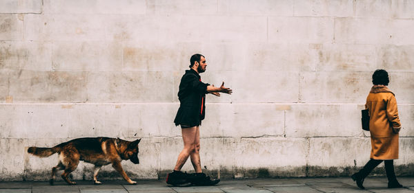 Side view of man with pants down walking by dog and woman against wall