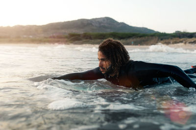 Serious surfer with curly hair in black wetsuit looking at coming wave while swimming on board in sea at sunset