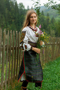 Girl with a bouquet of wildflowers stands on green grass near a wooden fence